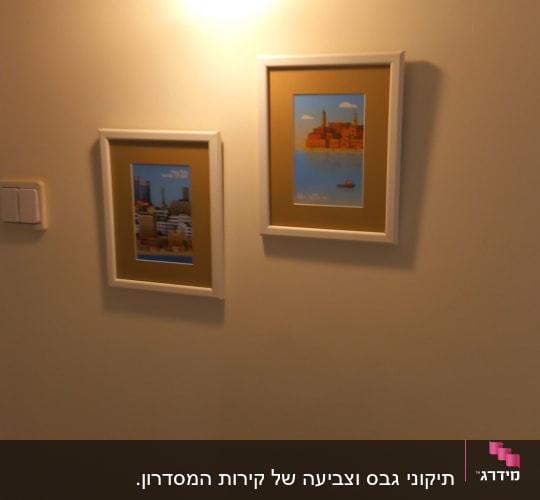 קיר פנים צבוע בגוון בהיר ועליו מנורת קיר ותמונות ממוסגרות